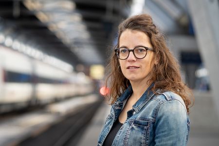 Rebekka Salm (Foto von Frederike Asael) Das Bild zeigt die Autorin Rebekka Salm, die auf einem Bahnhofperron steht. Salm trägt eine Brille und hat schulterlanges, leicht gewelltes hellbraunes Haar.